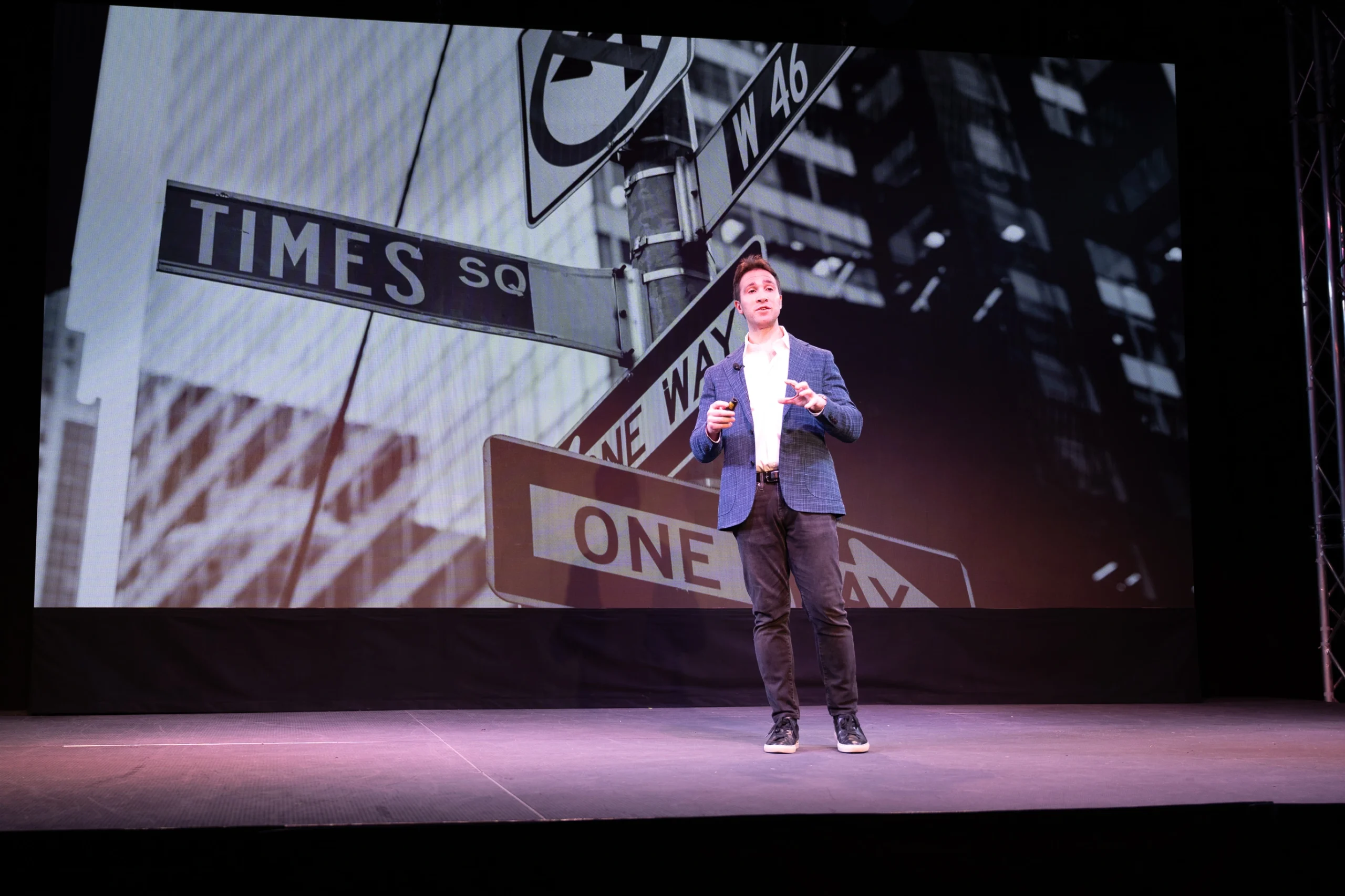 A man speaking on stage with a large city street sign image projected behind him.